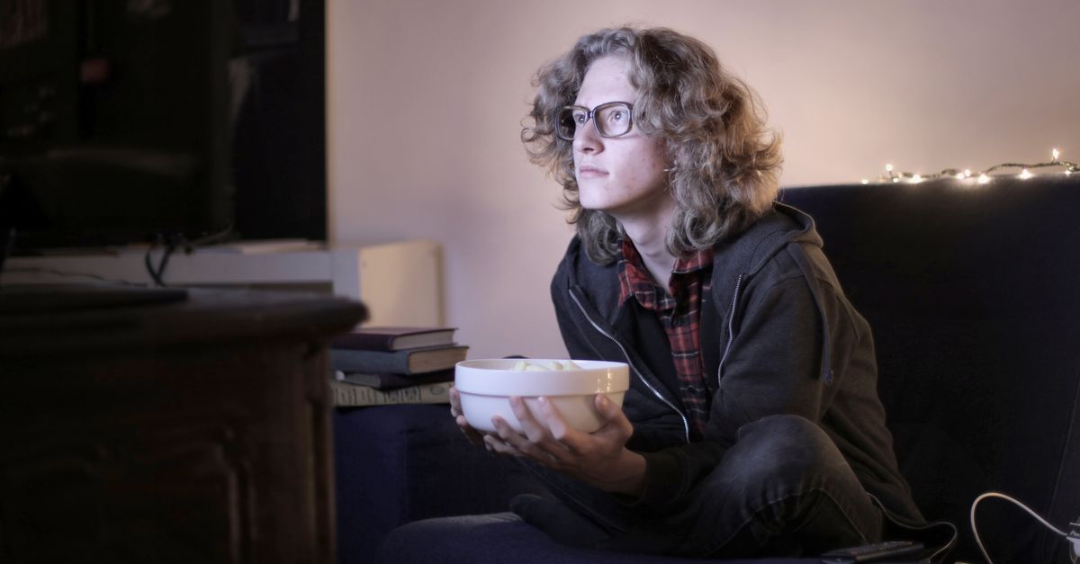 Adolescent concentré portant des lunettes, mangeant du popcorn devant la télévision dans un salon confortable.