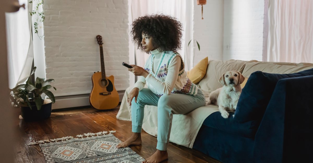 Jeune femme afro-américaine pieds nus sur un canapé, changeant de chaîne avec sa télécommande, chien allongé à côté